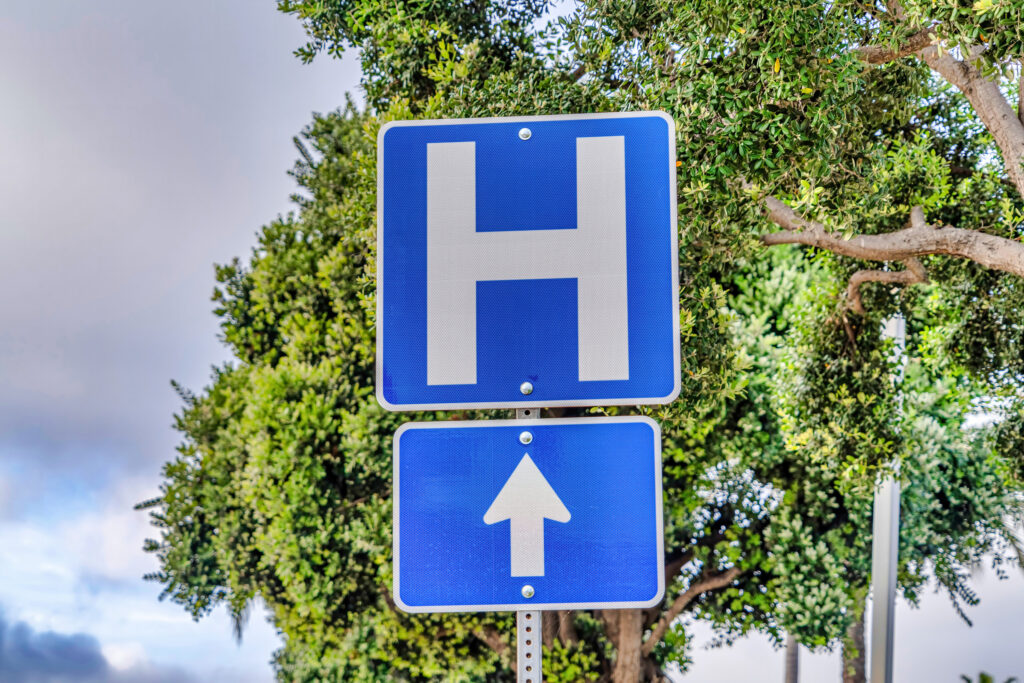 A sign with a capital H for hospital against trees and a blue sky