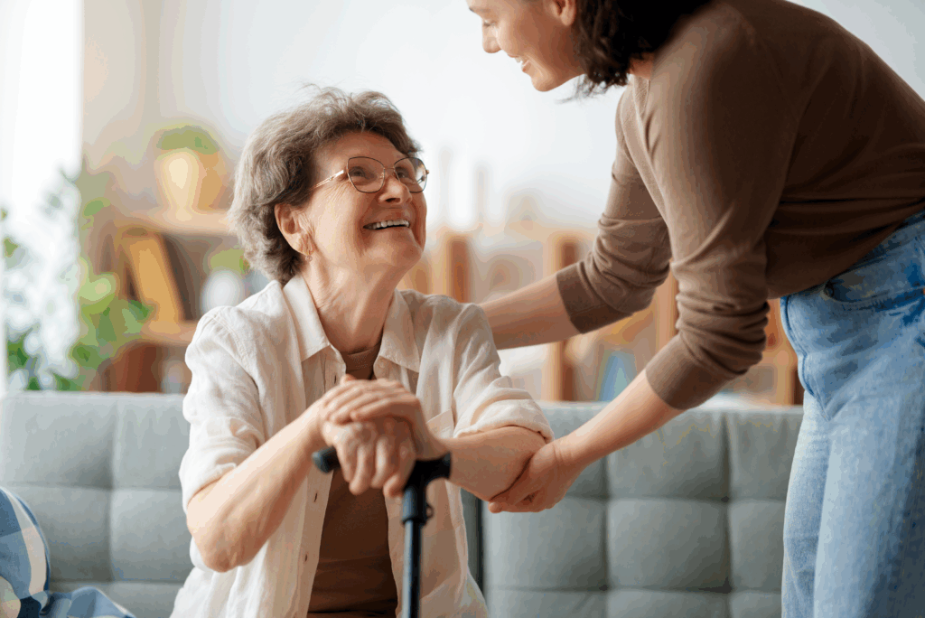 Elderly woman sits on a couch with a can in her hand and is looking up and smiling at a woman standing next to her with her hands on the lady's arm to help her get up.