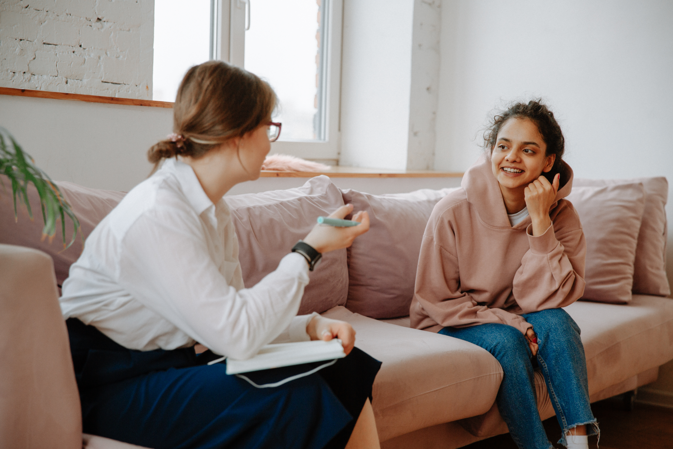 A woman wearing a white shirts sits on a beige couch across from another woman wearing a pink sweatshirt.